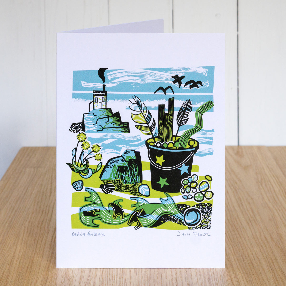 Beach Findings card. It depicts a beach with a bucket full of feathers, pebbles and driftwood. On the beach is seaweed, thrift and rocks.
