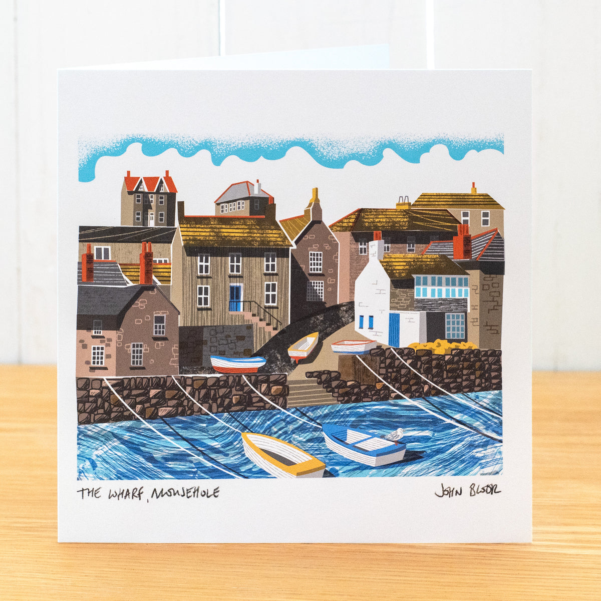 A greetings card showing houses on the harbour front of Mousehole, Cornwall. There are also boats on the road leading down to the harbour and boats on the water.