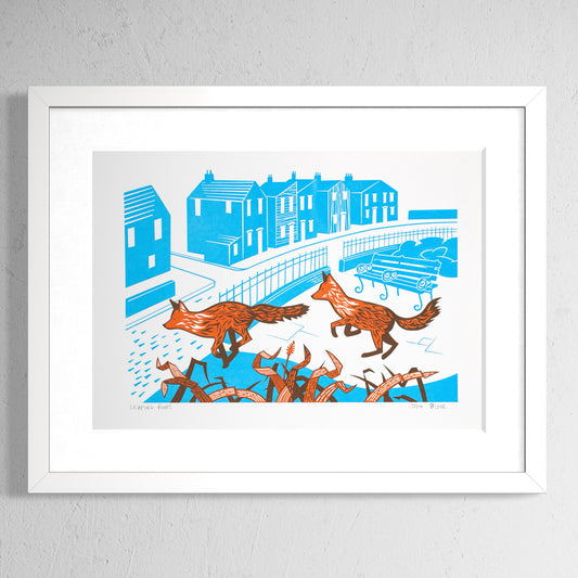 Leaping Foxes screenprint in orange and cyan showing two foxes leaping over grasses in front of an urban street scene. Shown in a white frame.