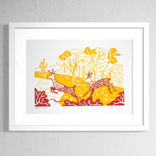 Leaping Stags screenprint in orange and red showing two stags leaping over bushes in front of woods. Shown in a white frame.