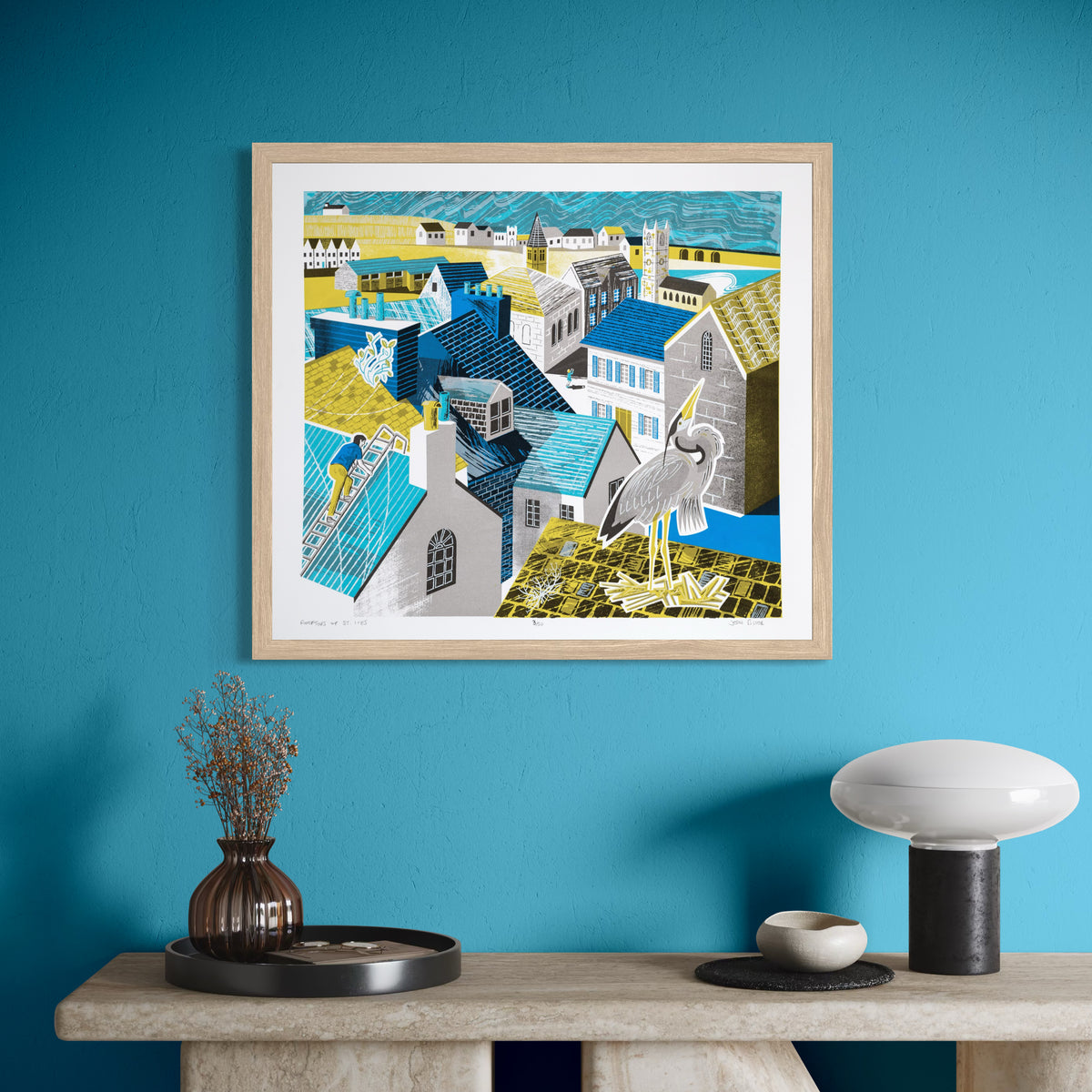 Rooftops of St. Ives screenprint featuring the rooftops of St. Ives, a heron in the foreground nesting on a roof, a man up a ladder repairing a roof and a tourist taking a photograph. Shown in an oak frame on a turquoise wall.