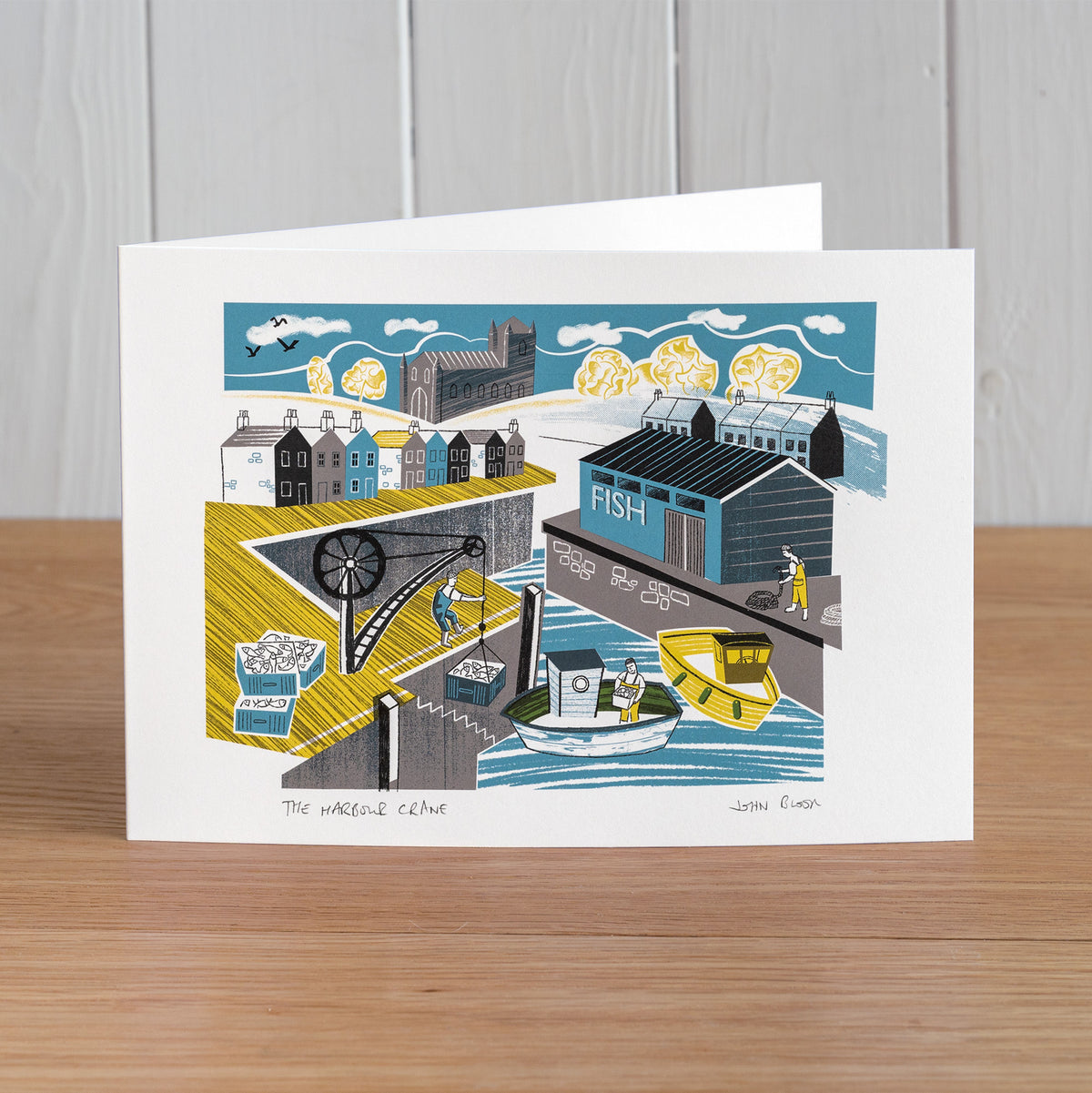 The Harbour Crane greetings card featuring fishermen working on a harbour, unloading a boat using a crane. There's a warehouse and houses behind.