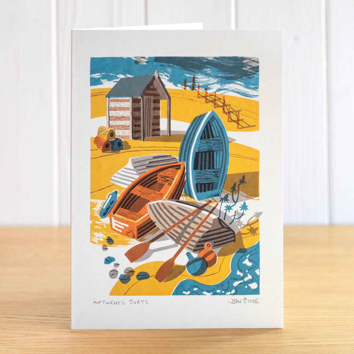 Upturned Boats greetings card, depicting four small wooden boats on a beach, some upturned, with a hut behind that.