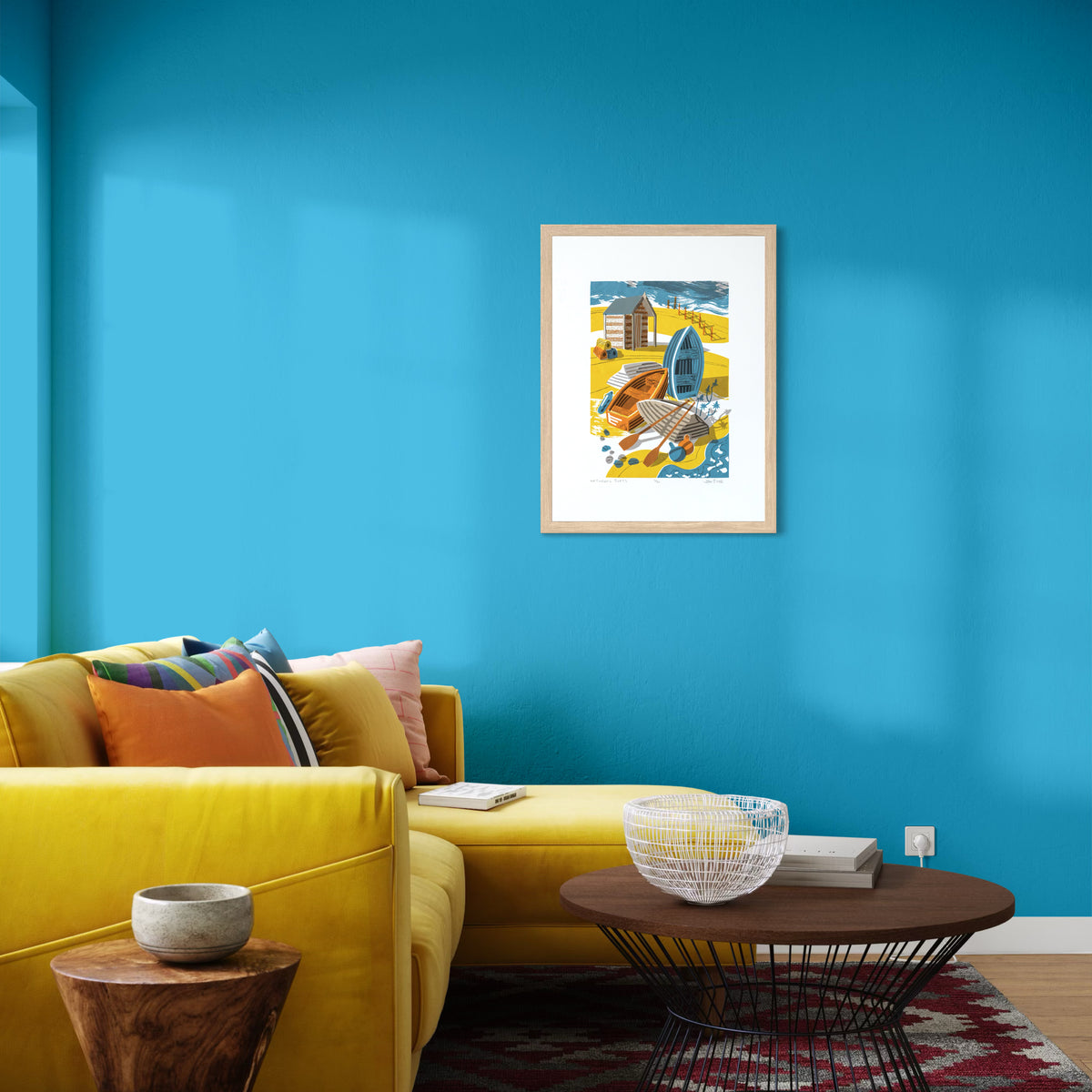 Upturned Boats screenprint, depicting four small wooden boats on a beach, some upturned, with a hut behind that, buoys and a fence. Shown on a blue wall in a living room.