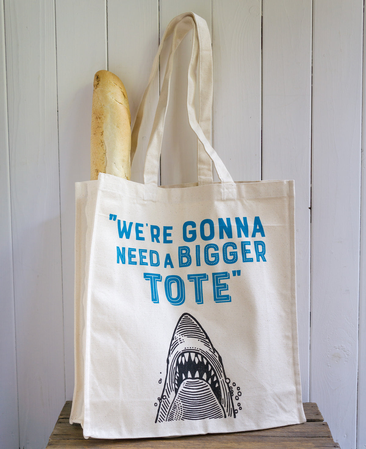 A high quality canvas shopper bag featuring a shark design and the words We're Gonna Need a Bigger Tote. Showing the bag holding a baguette.
