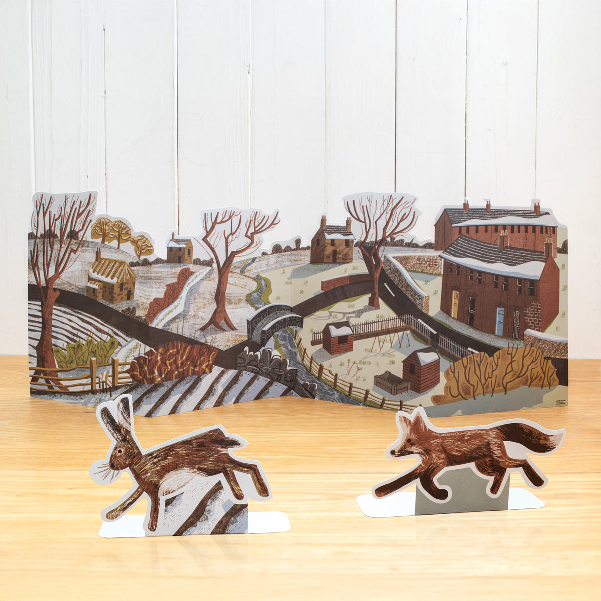 This is a stand-up, concertina fold, Winter scene. It features an urban moving into a rural scene with a river, allotment, trees, houses, sheds and hedgerows.

In front are two separate stand up animals; a fox and a hare.