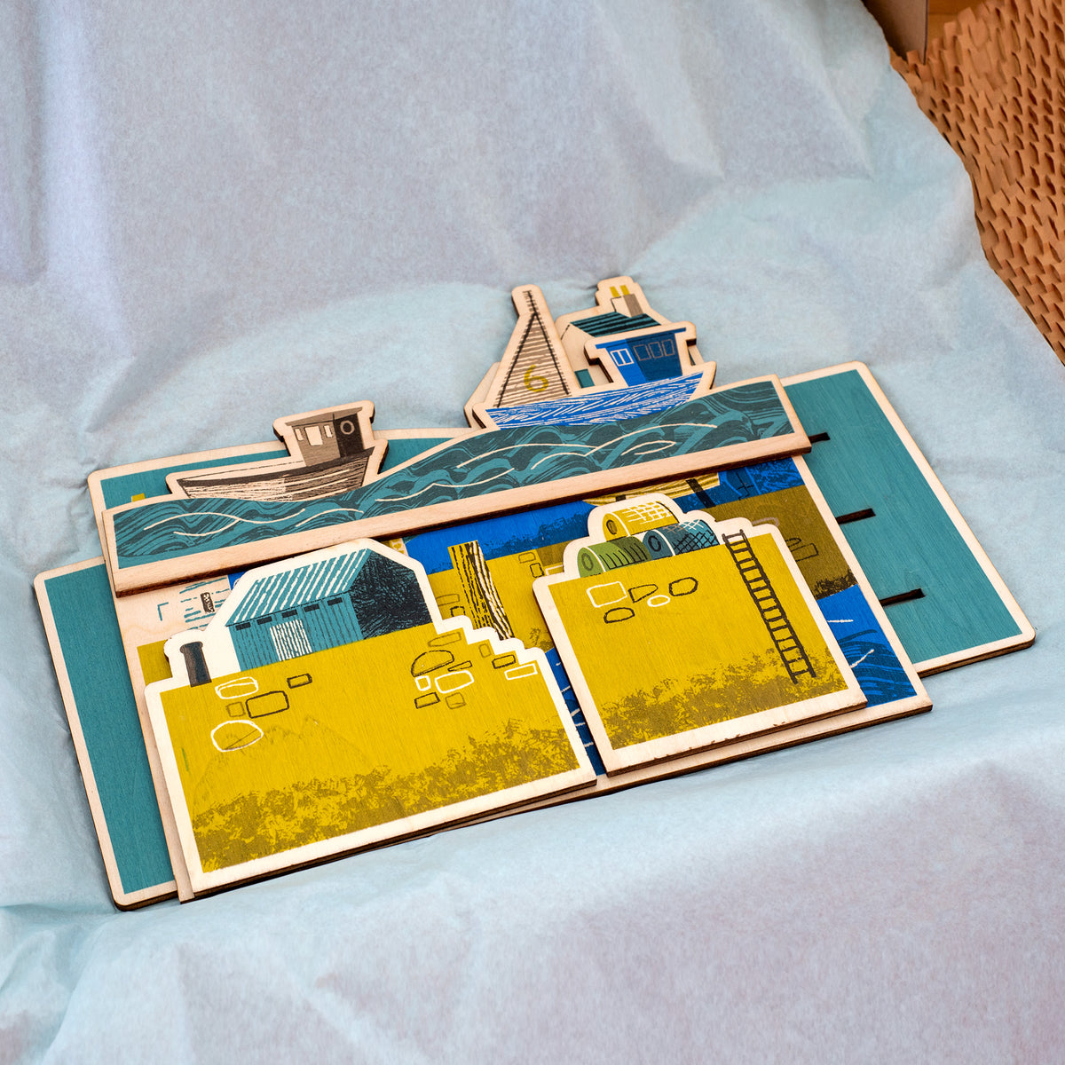 Wooden Harbour hand printed scene. It shows a harbour scene with sea and boats in the foreground, a harbour wall with ladder, lobster pots and a hut, behind is a fisherman working on a boat and houses.