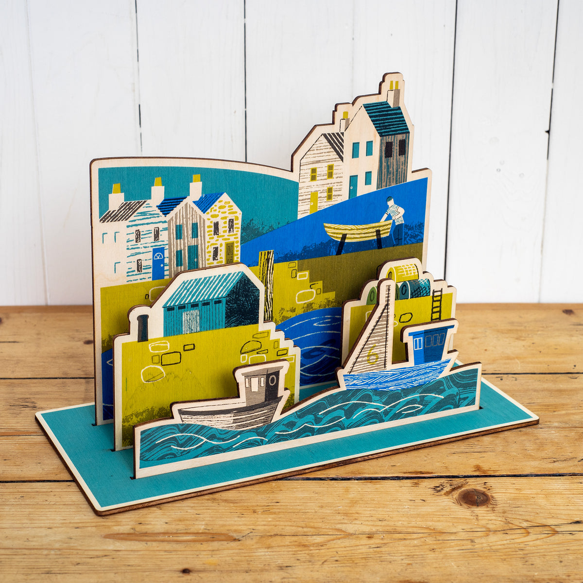 Wooden Harbour hand printed scene. It shows a harbour scene with sea and boats in the foreground, a harbour wall with ladder, lobster pots and a hut, behind is a fisherman working on a boat and houses.