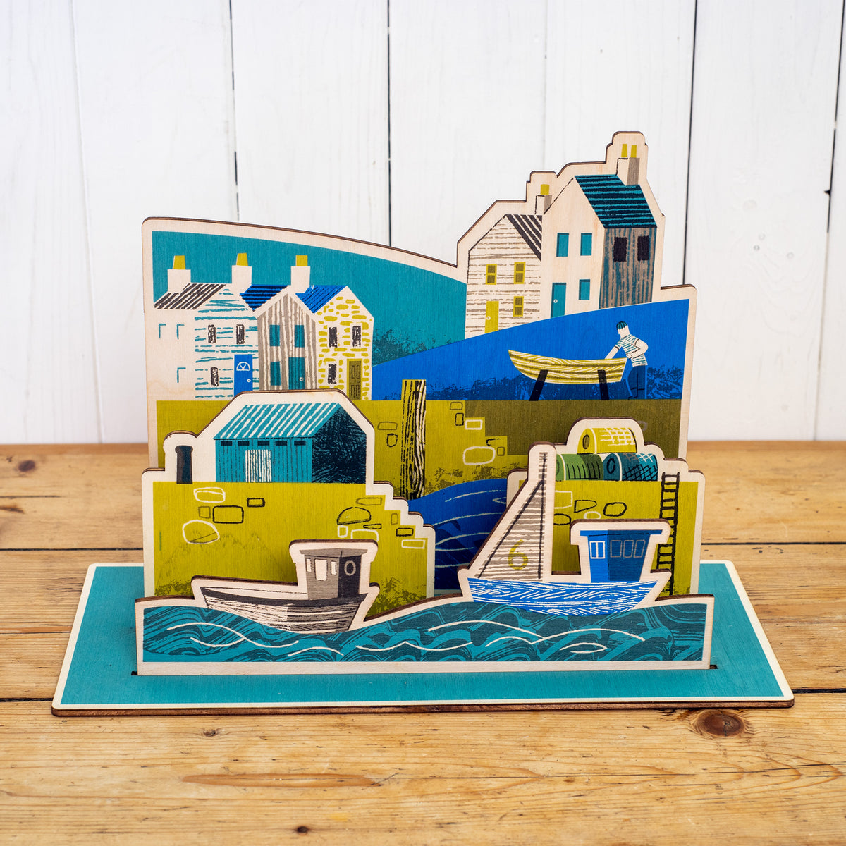Wooden Harbour hand printed scene. It shows a harbour scene with sea and boats in the foreground, a harbour wall with ladder, lobster pots and a hut, behind is a fisherman working on a boat and houses.