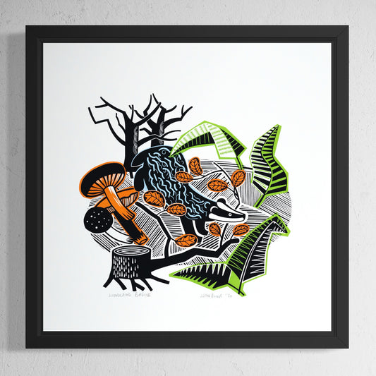 Woodland Badger hand printed screenprint. It depicts a badger walking amongst fallen leaves, mushrooms, tree trunk, ferns and trees. In a black frame.