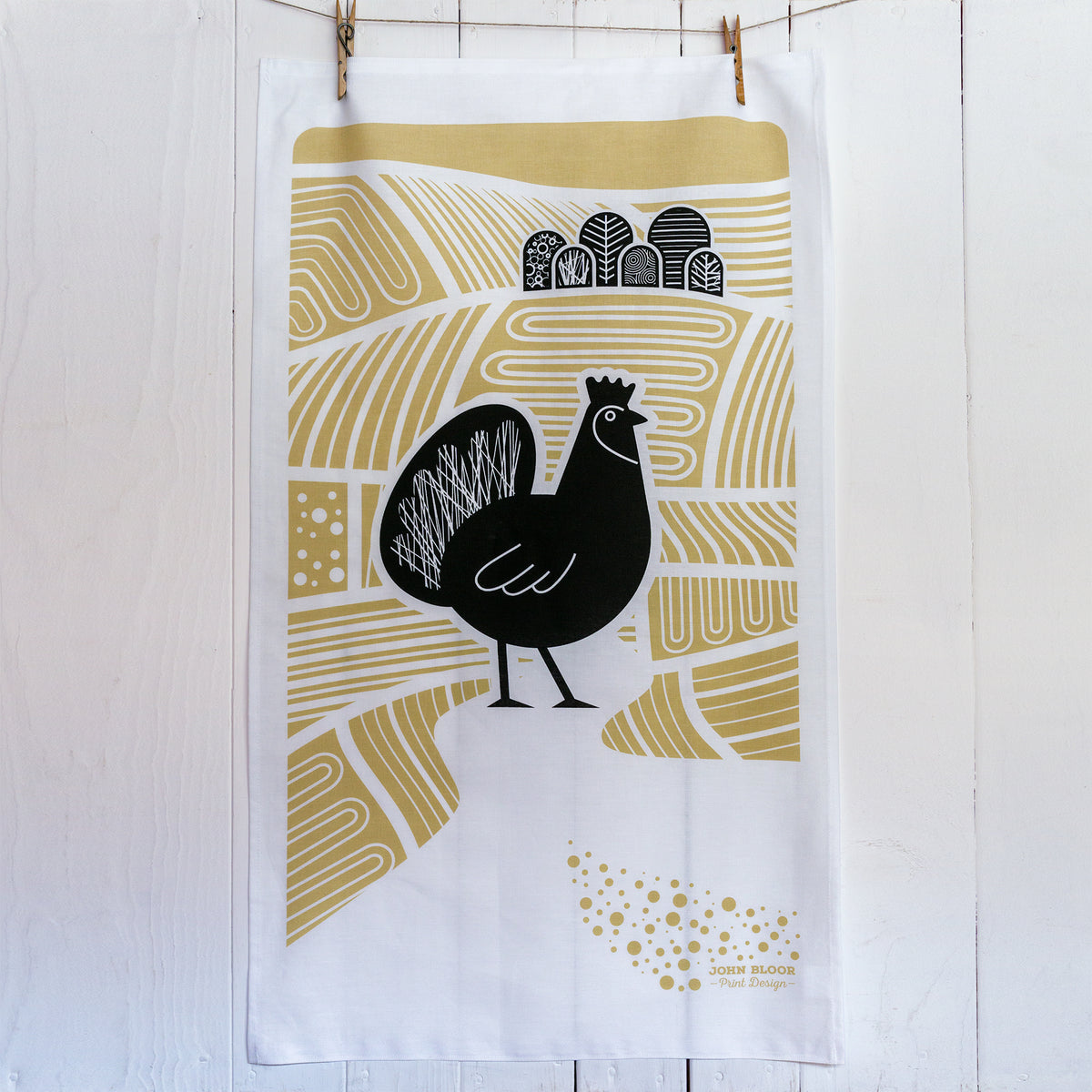 A screen printed tea towel in black and brown featuring a chicken standing amongst ploughed fields and trees.