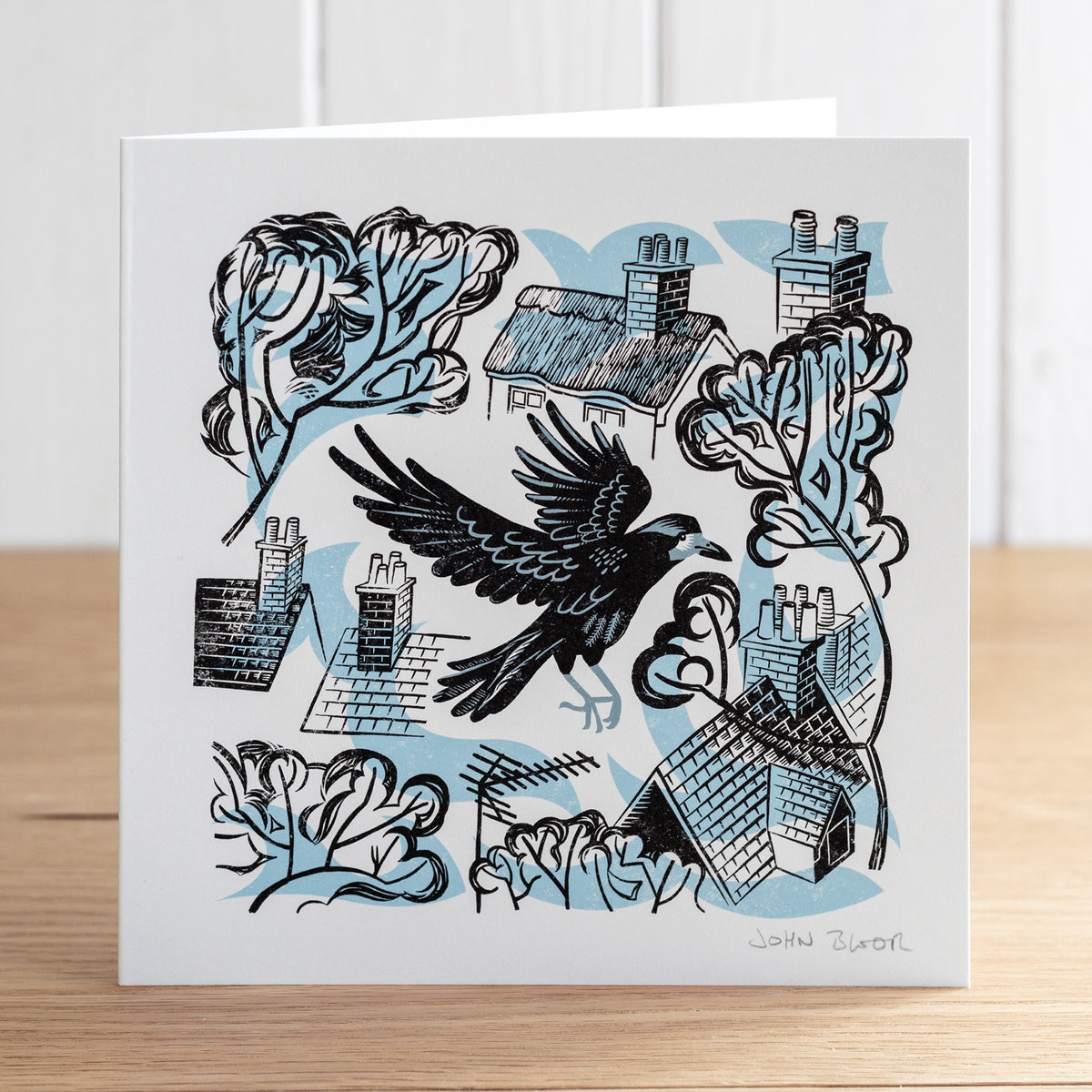 Take Flight Crow greetings card. This card depicts a crow flying between rooftops and trees. 